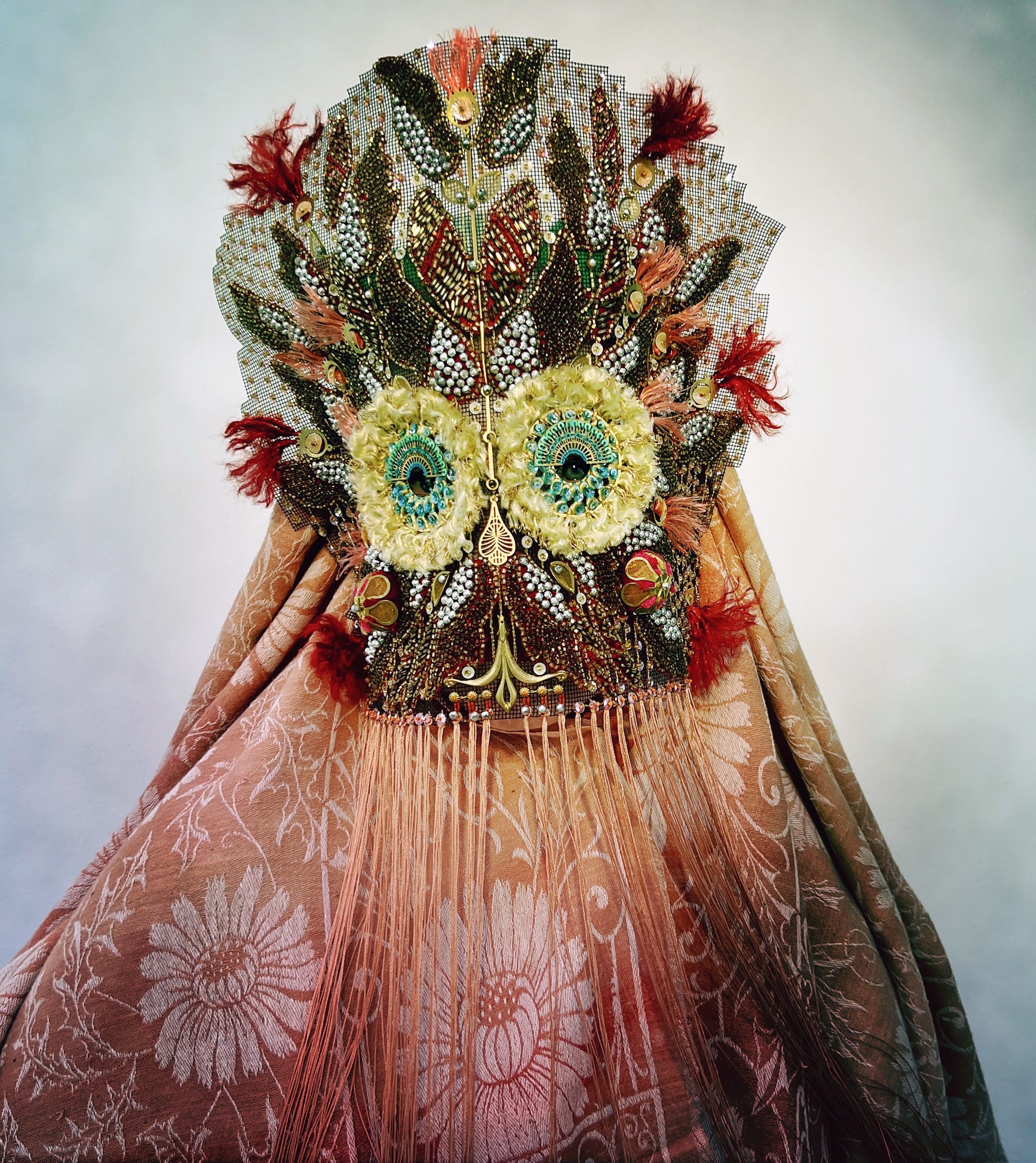 an ornate mask by Magnhild Kennedy with owl-like eyes, feathers, beads, and a patterned peach textile covering the body