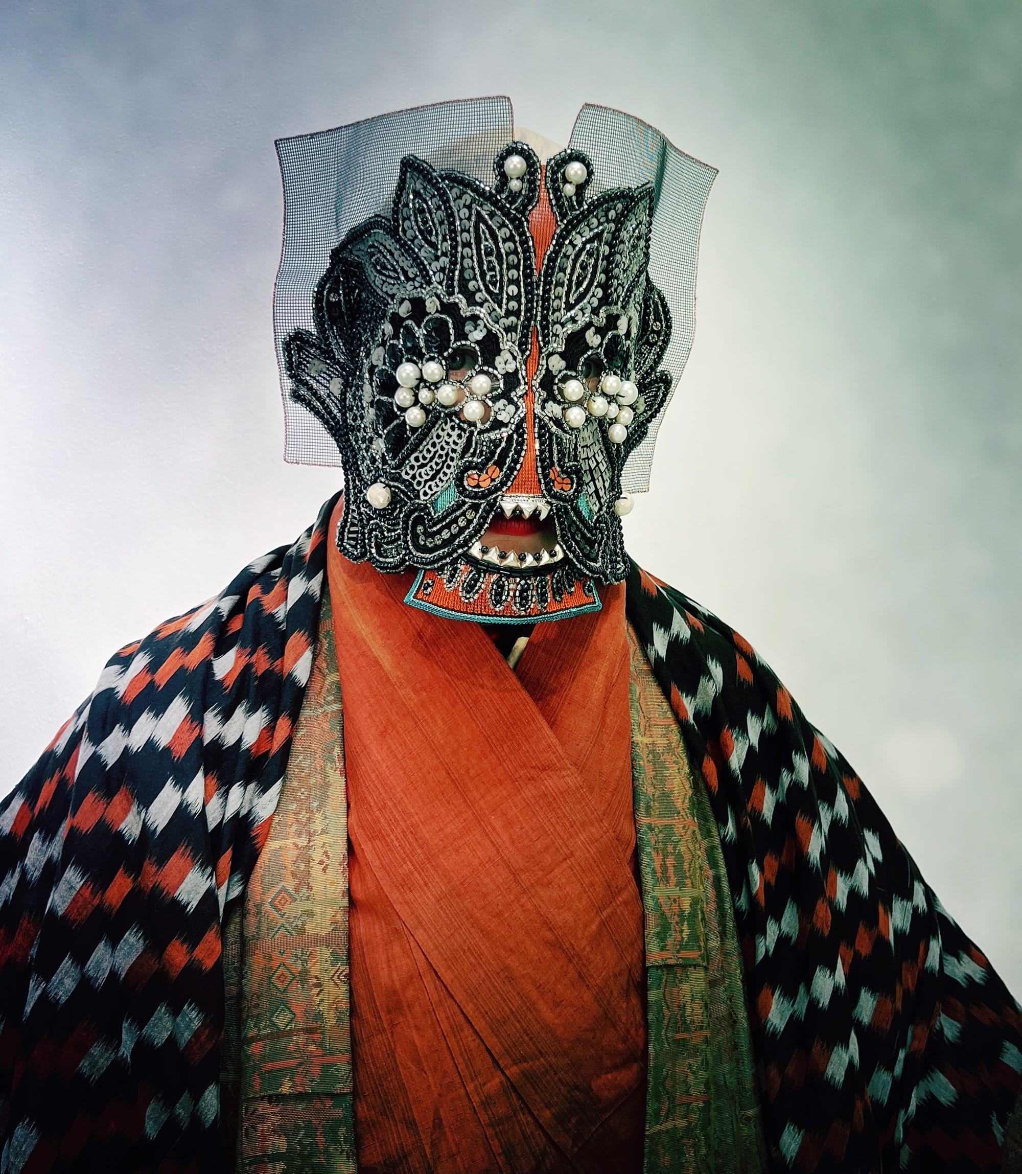 an ornate mask by Magnhild Kennedy with black and white beads, and mesh, and a patterned red and black textile covering the body