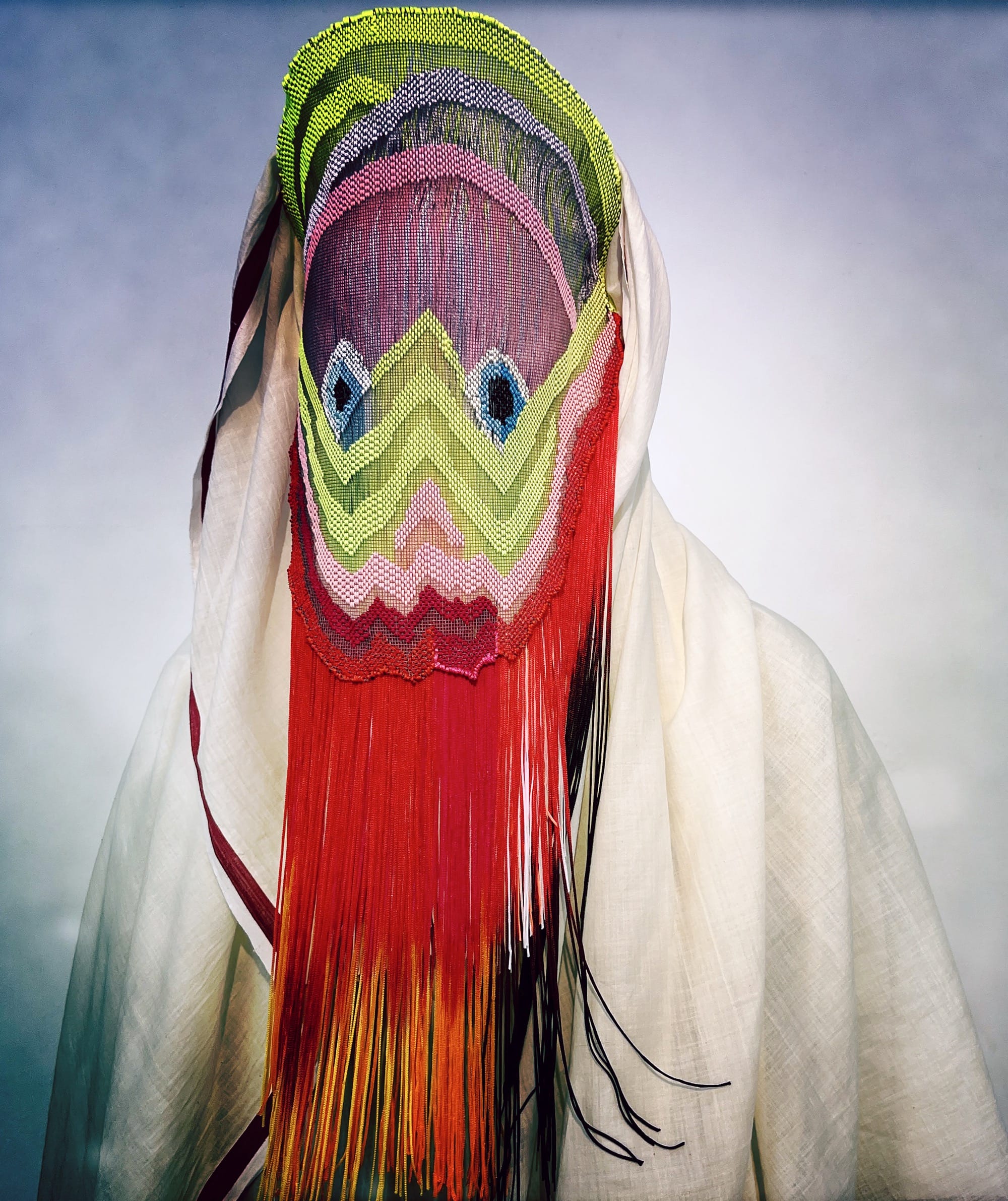 an ornate mask by Magnhild Kennedy with long draping red, purple, green, and blue fringe and a white textile covering the body