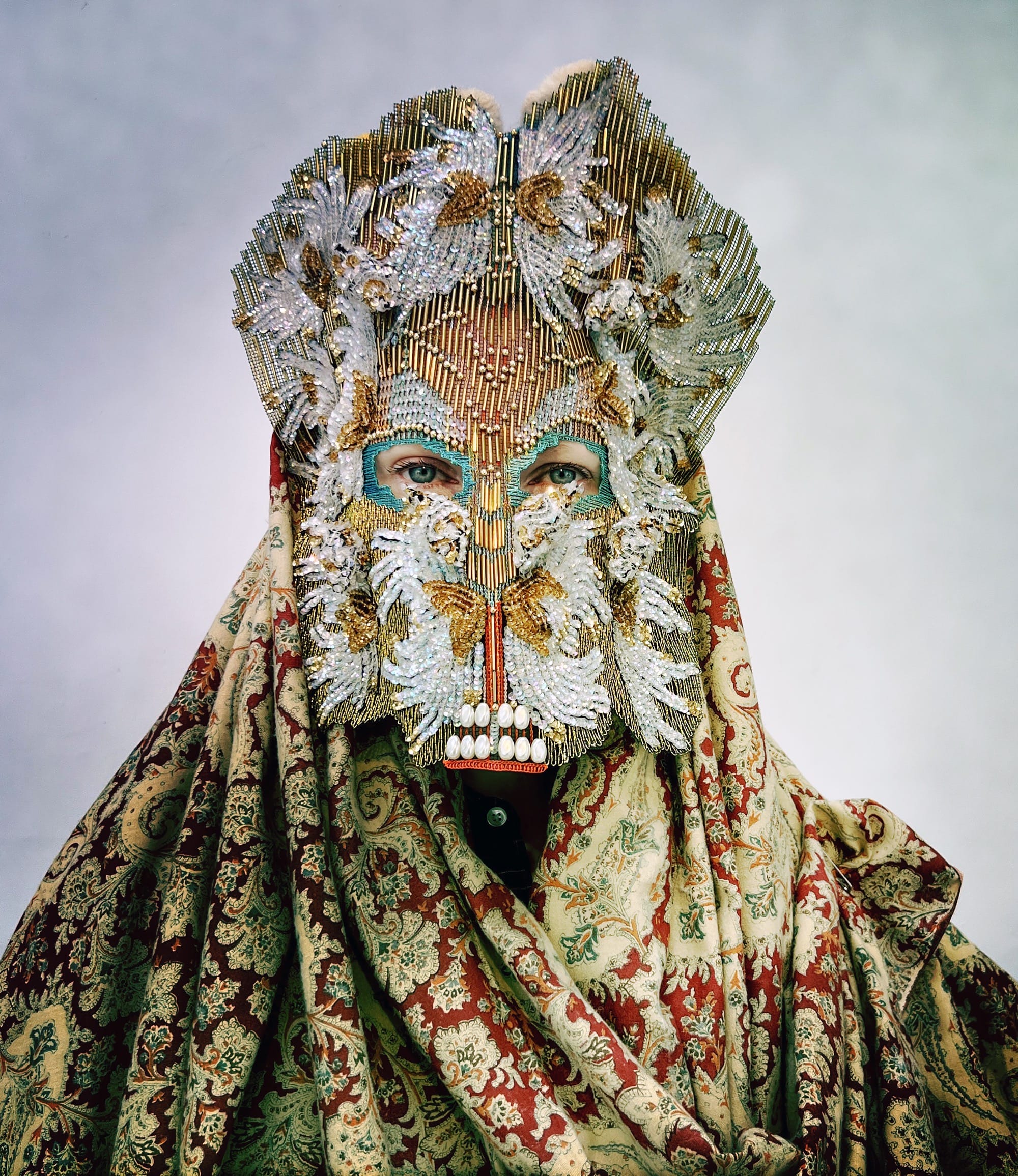 an ornate mask by Magnhild Kennedy with white and gold beads and a patterned gold and red textile covering the body